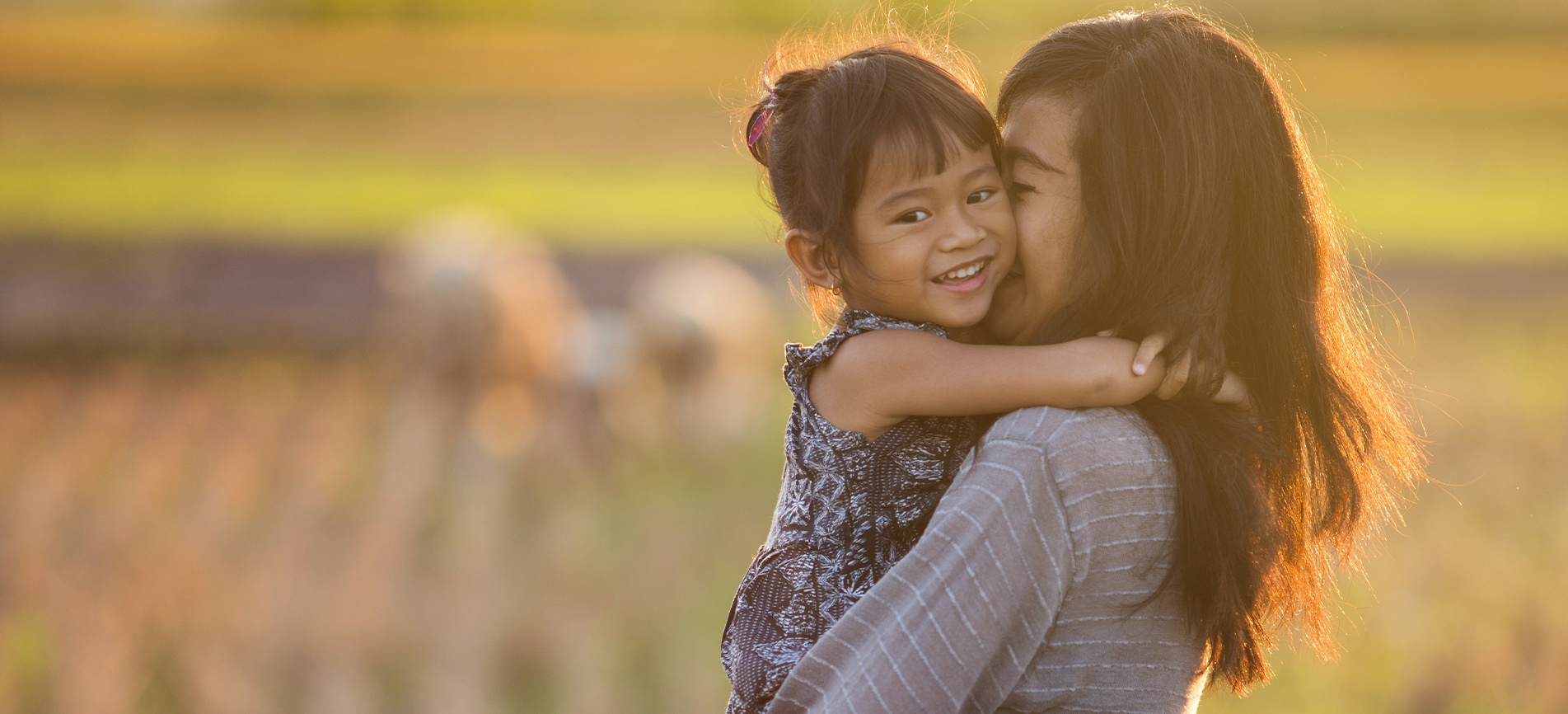 An Asian mother holds her smiling young daughter as they look out over farmland in late afternoon sun