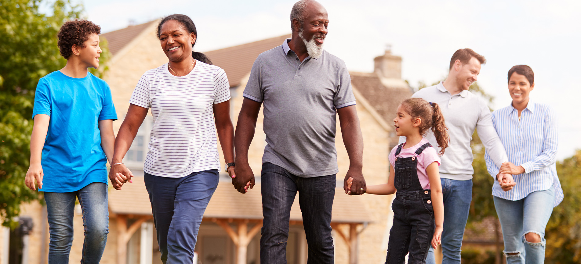 A multi-ethnic and multi-generational family walks hand-in-hand outside of an older home on a sunny day