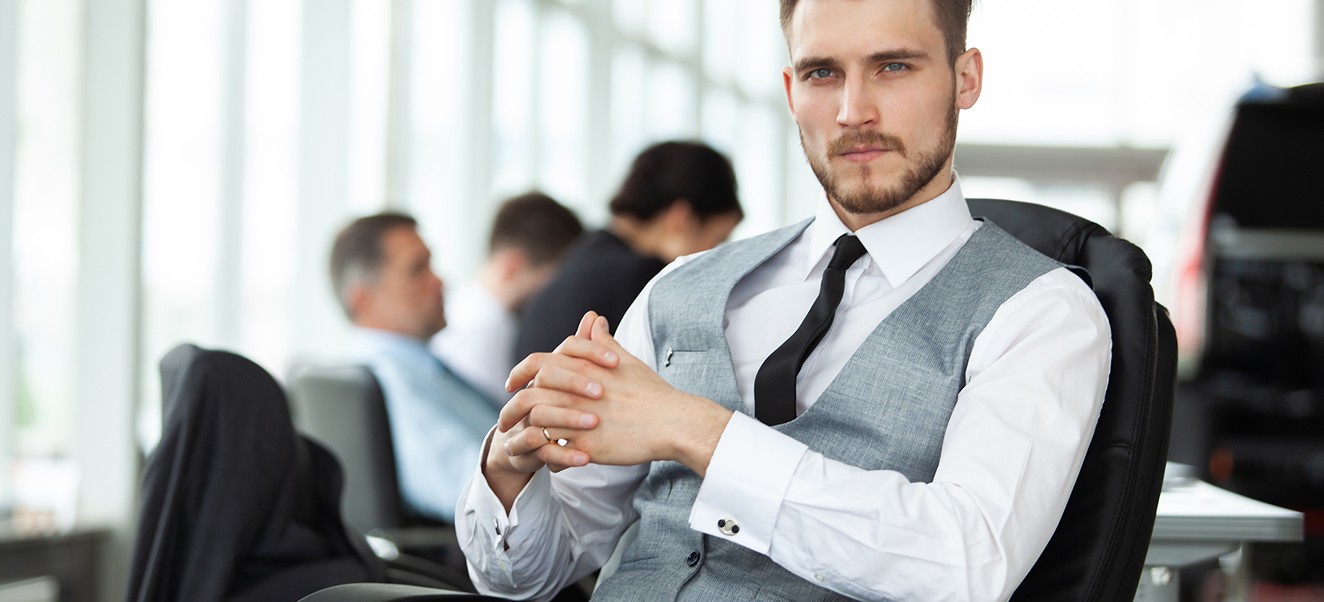 A well-dressed young business man sits in a bright conference room with colleagues in the background