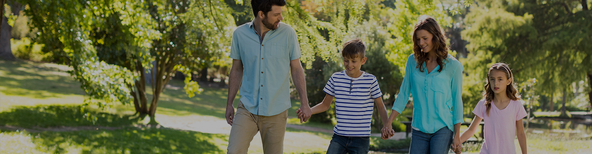A young family with their children hold hands as they walk through a sunny city park filled with trees