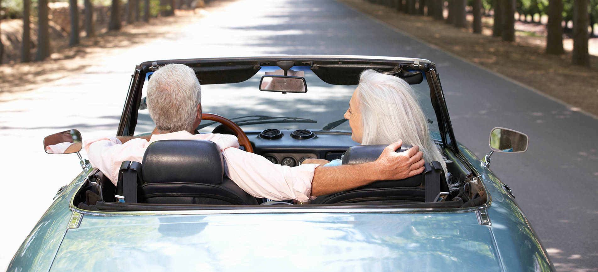 A happy older couple drive in their convertible down a tree-lined street.