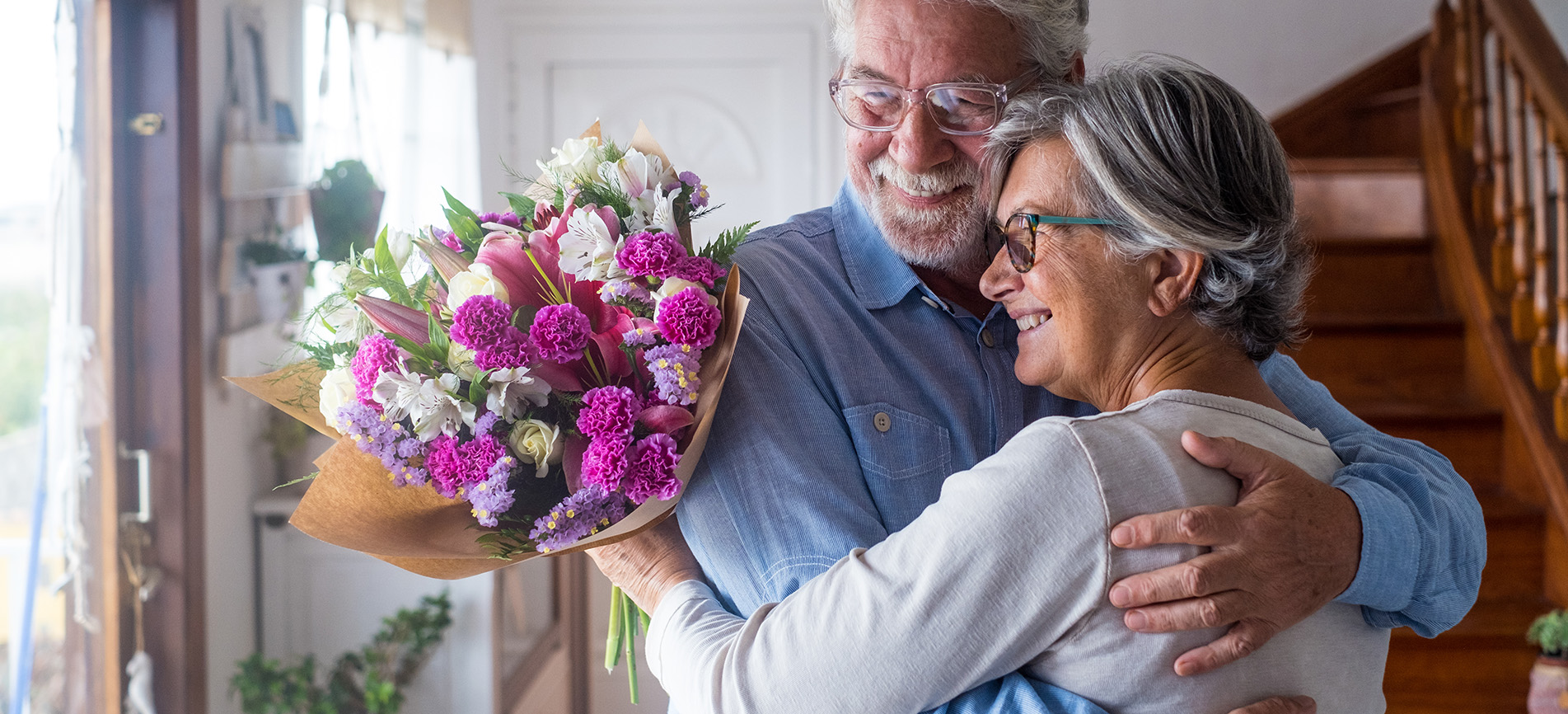 An older man presents an older woman a bouquet of flowers as they embrace in a brightly-lit room.