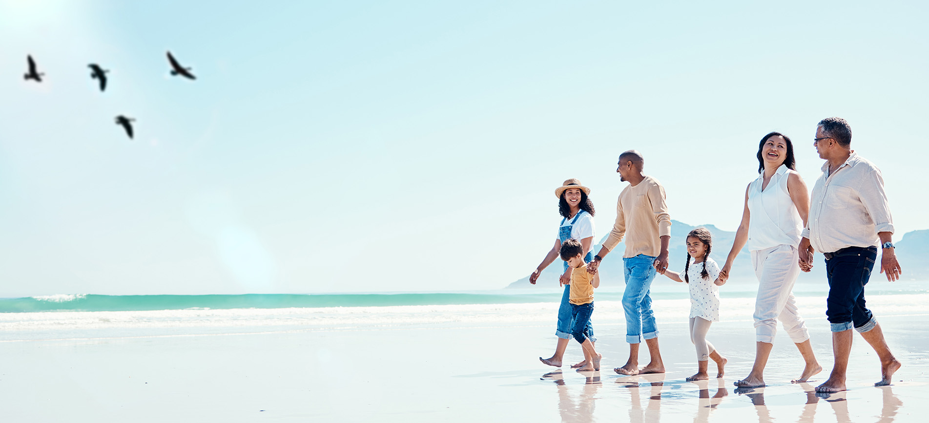 A family with their children and parents walks along ocean on the beach on a bright sunny day with birds flying overhead.