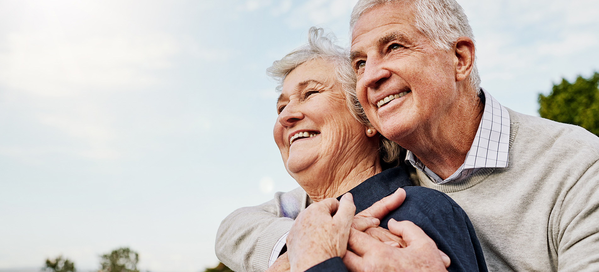 A happy older couple are shown in an embrace against a partly cloudy blue sky.