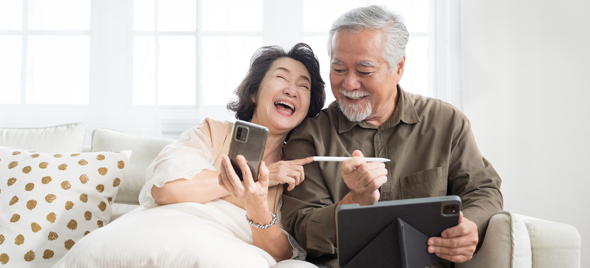 An older couple laugh and enjoy their time together while using a smartphone and tablet computer, relaxing on a couch in a sunny room in their home.