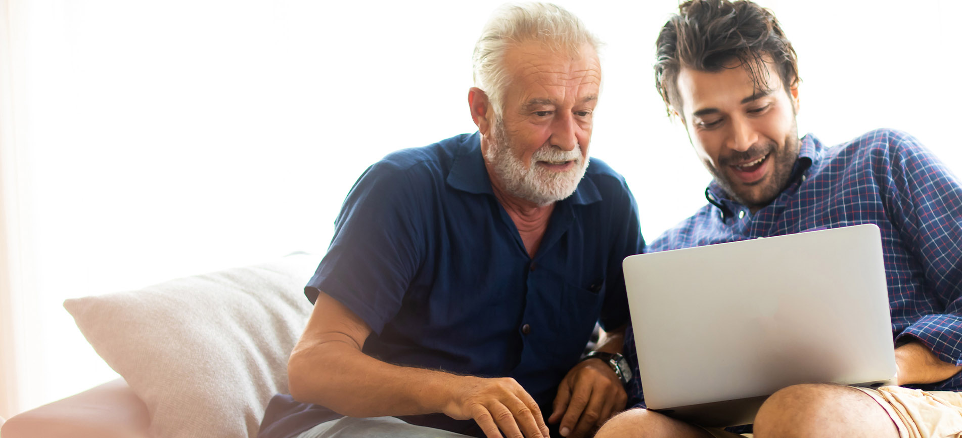 A father and son excitedly work at a laptop computer, sitting on a couch against a bright background.