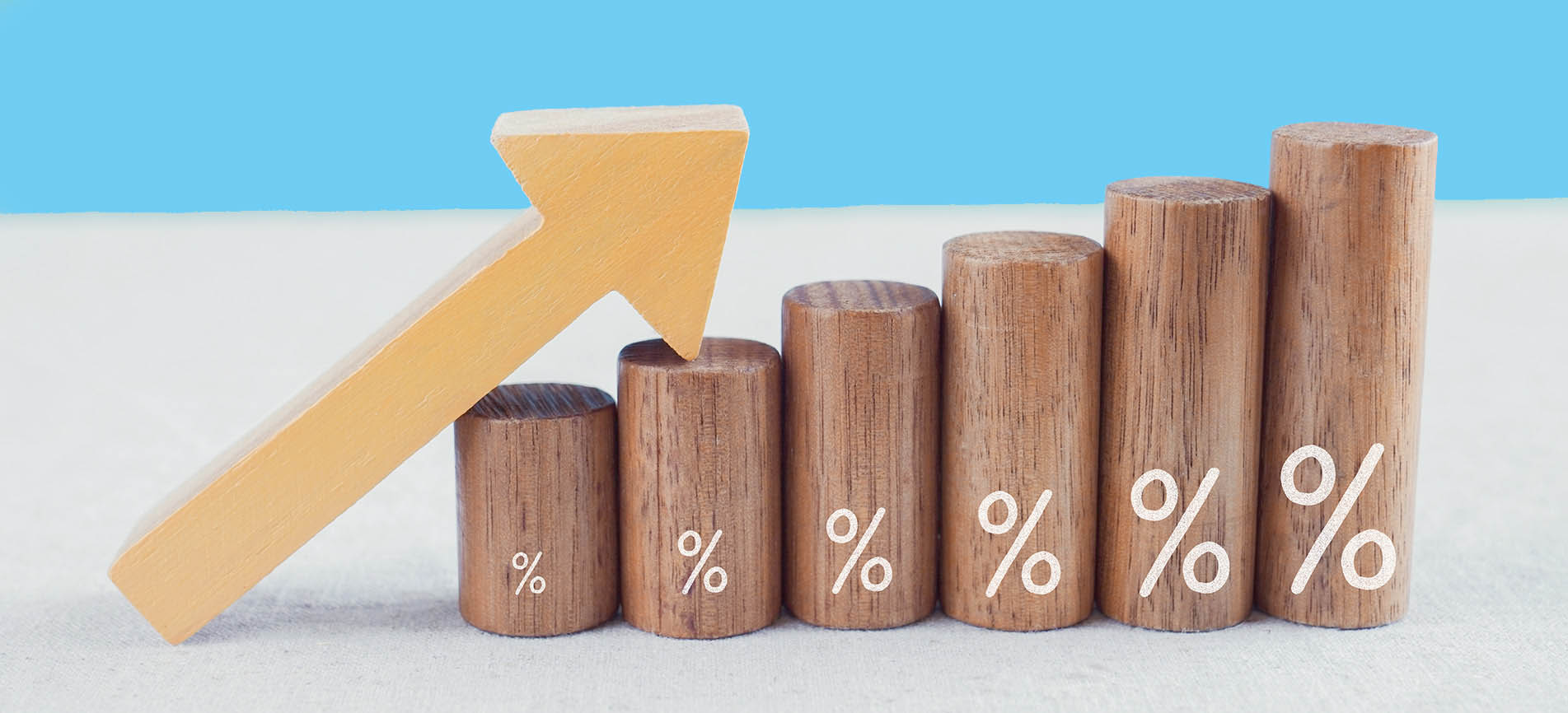 A wooden, pale-yellow, long arrow points up and to the right, leaning on against a set of round wooden posts with percent signs on them, on a pale blue and light gray background.