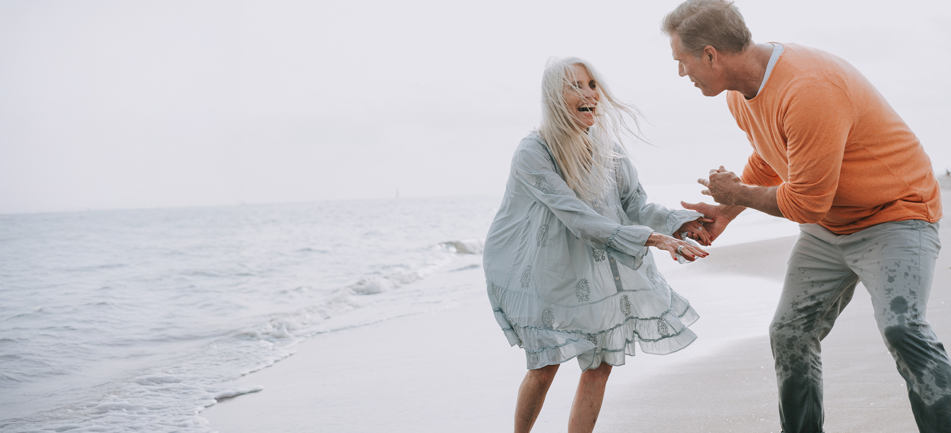An older couple happily walks along the beach with light ocean waves in the background.