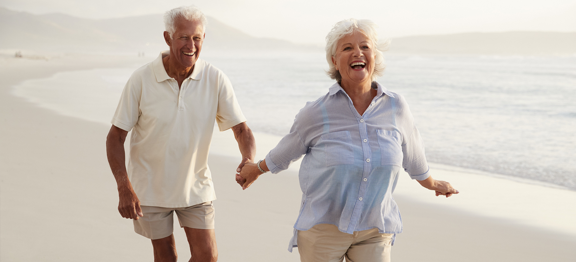 An happy elderly couple hold hands and walk on the beach on a bright day