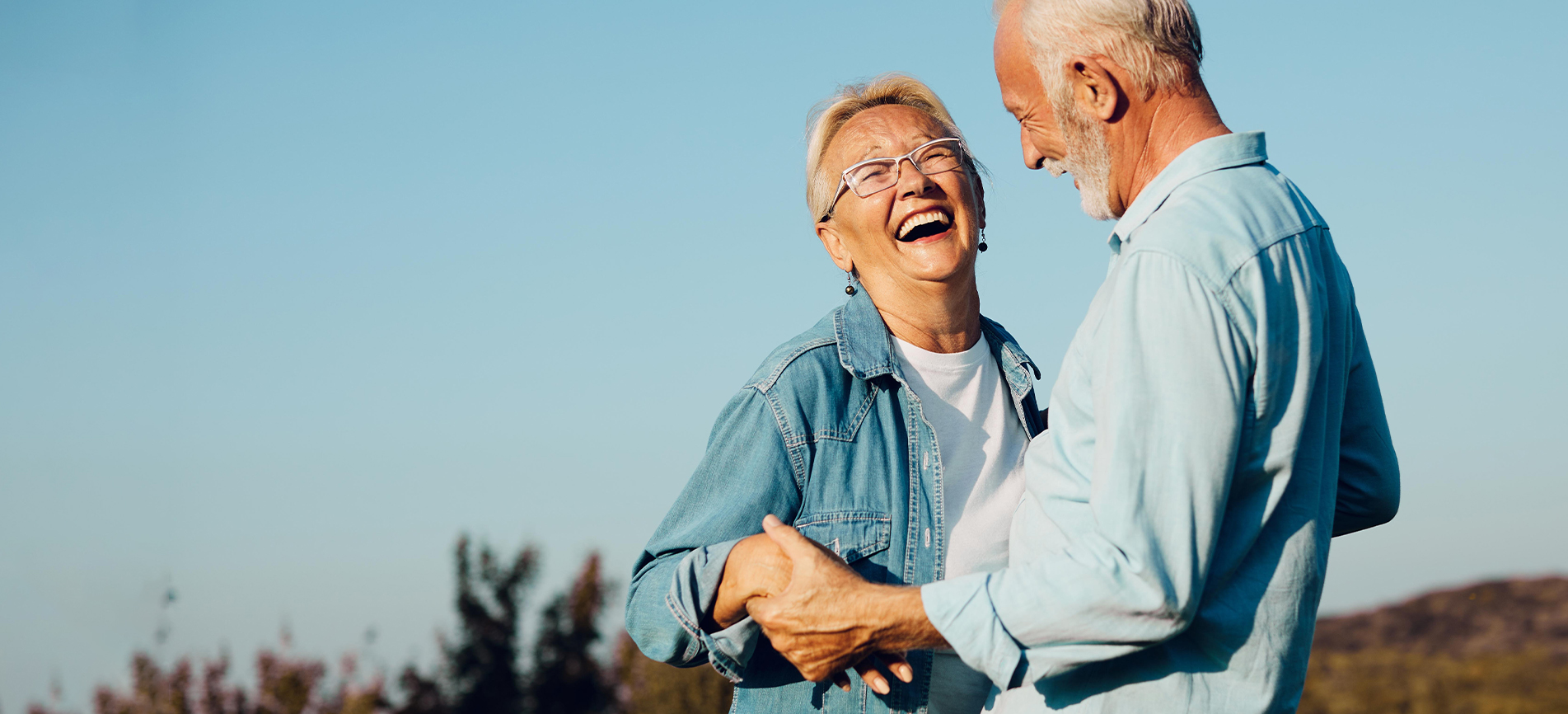 An excited, happy older couple holds hands and dances along the beach against a beautiful blue sky.