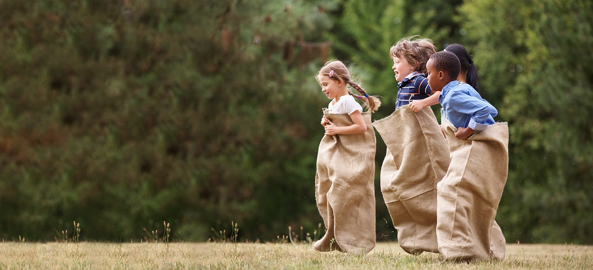 Young children in sacks prepare to run a race in a lush green park on a sunny day
