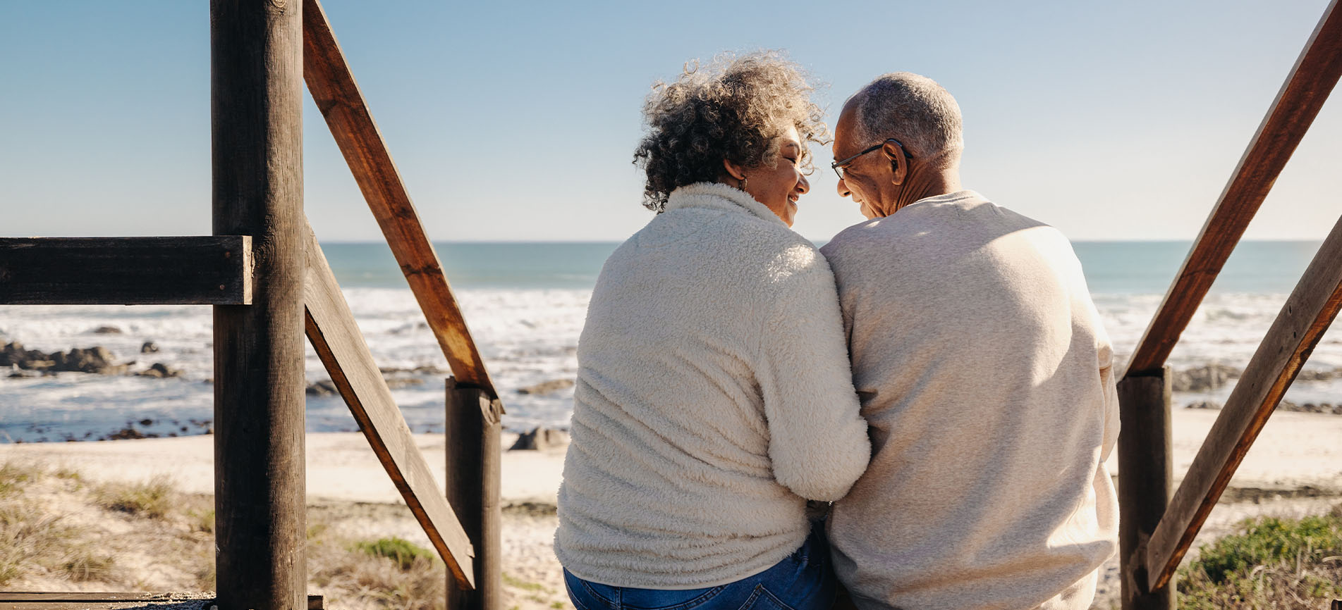 An older couple smile at each other while sitting on a staircase at the beach looking at the ocean.